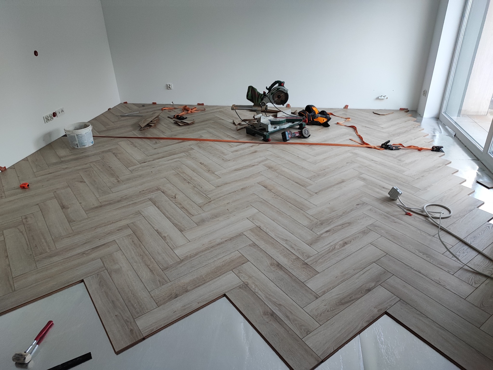Układanie parkietu w jodełkę w jasnym wnętrzu. Widoczne narzędzia, listwy przypodłogowe i częściowo ułożona podłoga. Etap prac wykończeniowych z bliska.