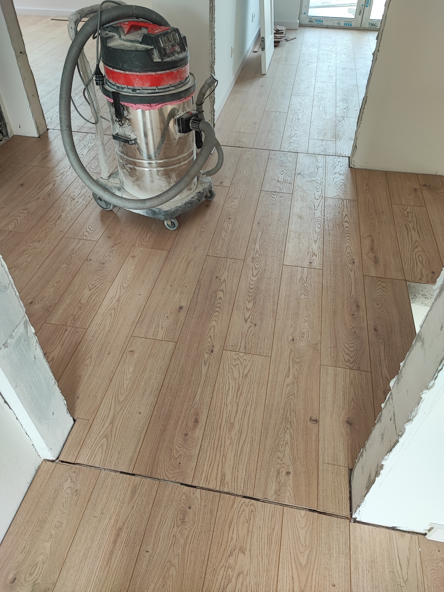 Ułożona podłoga z paneli w odcieniach brązu, widoczny odkurzacz przemysłowy i fragmenty ścian w trakcie wykończenia. Perspektywa z góry, wnętrze w trakcie prac.