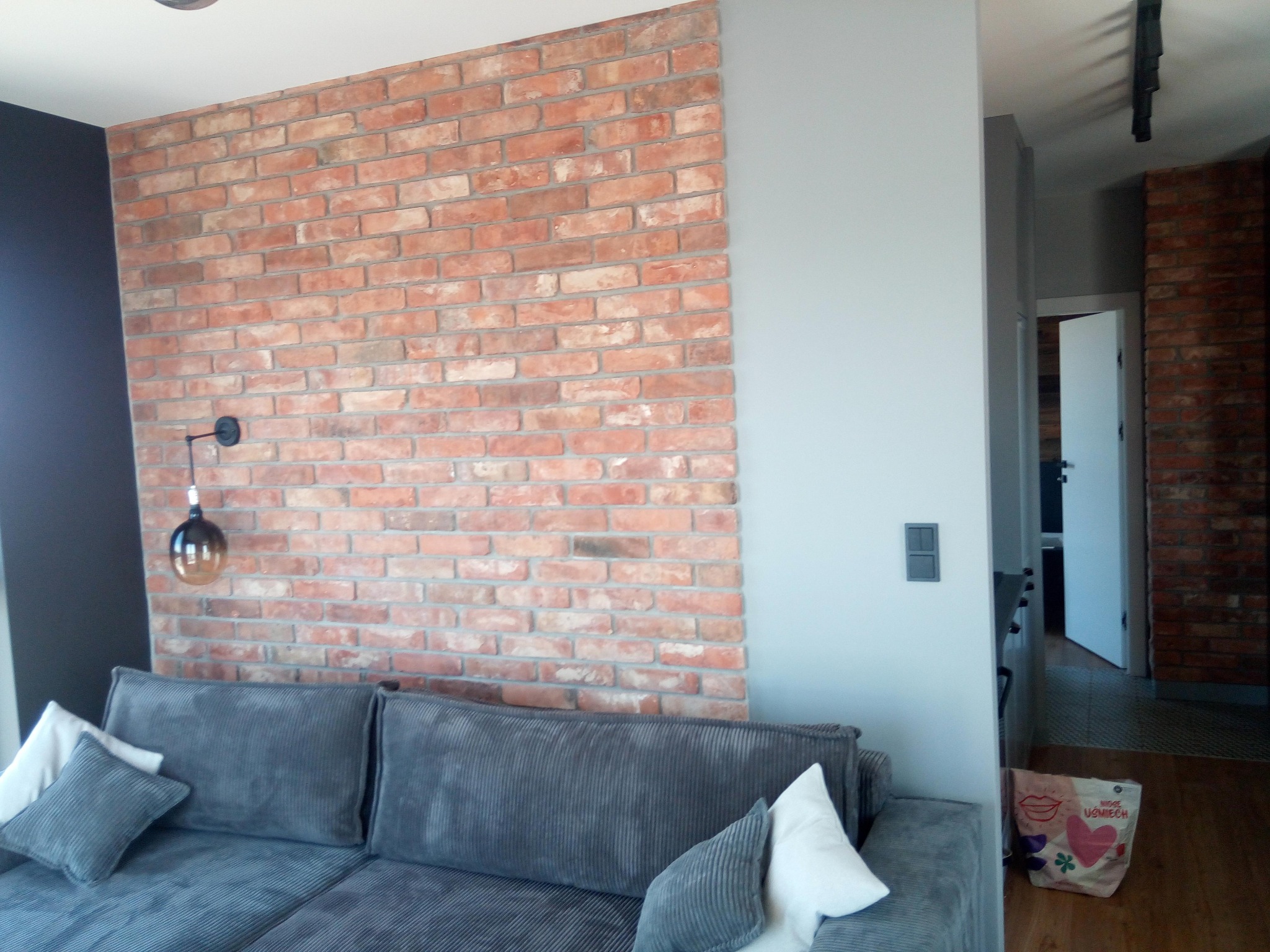 Ceglana ściana w salonie z szarą sofą i lampą wiszącą. Widoczne detale wykończenia, takie jak kontakt i torba na podłodze. W tle otwarte drzwi do innego pomieszczenia.