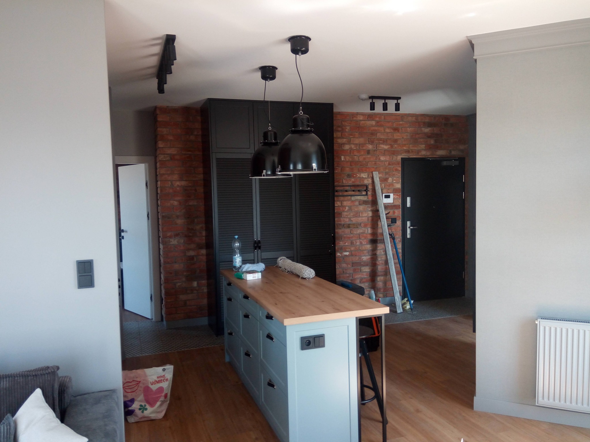 Loftowe wnętrze po remoncie: ceglana ściana, wyspa kuchenna z drewnianym blatem, czarne lampy wiszące. Widoczne detale wykończenia i oświetlenie.