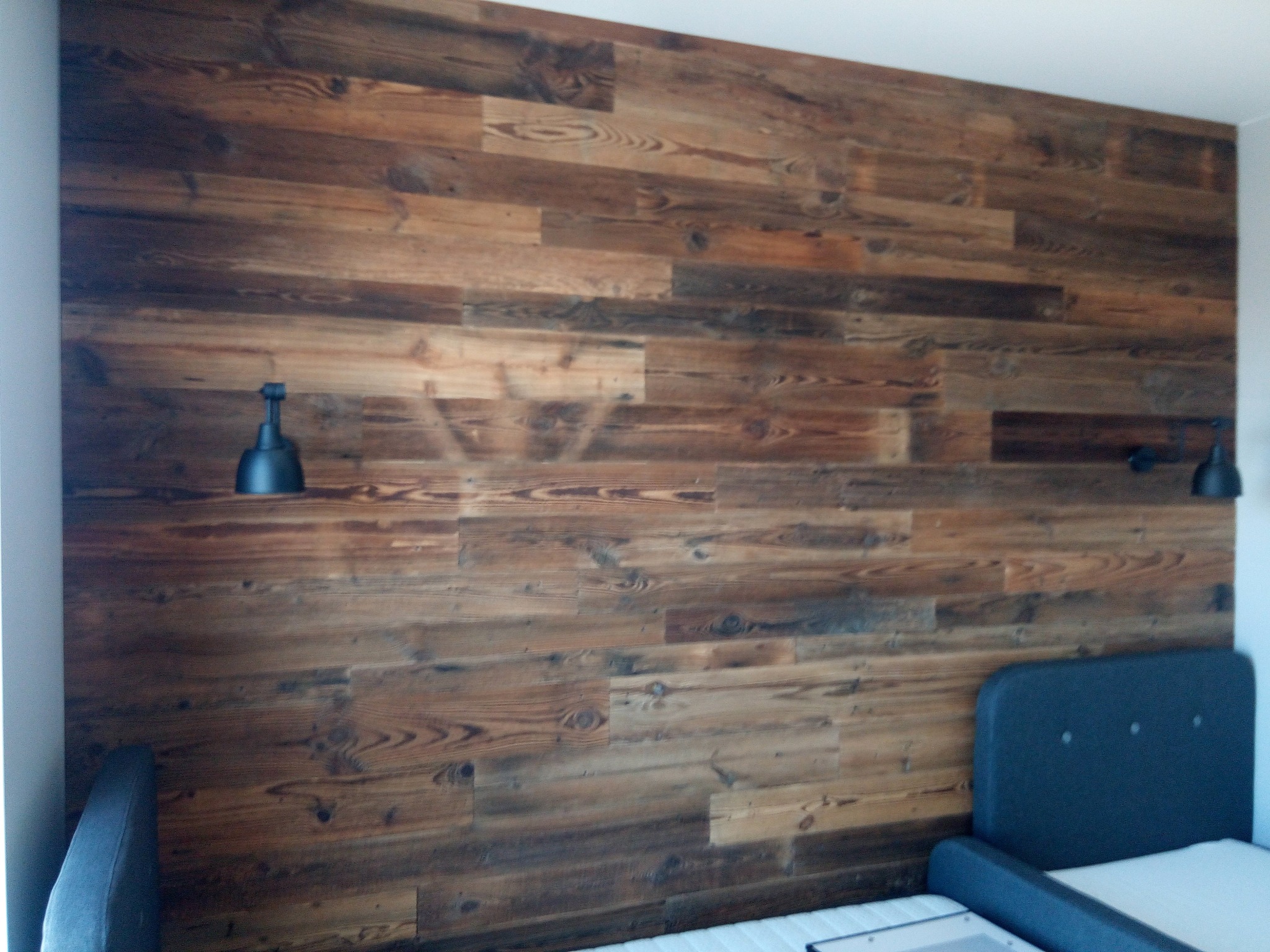 Ściana sypialni wykończona panelami z ciemnego drewna, z widocznymi sękami i naturalnym usłojeniem. Dwie czarne lampki nocne zamontowane po bokach łóżka.