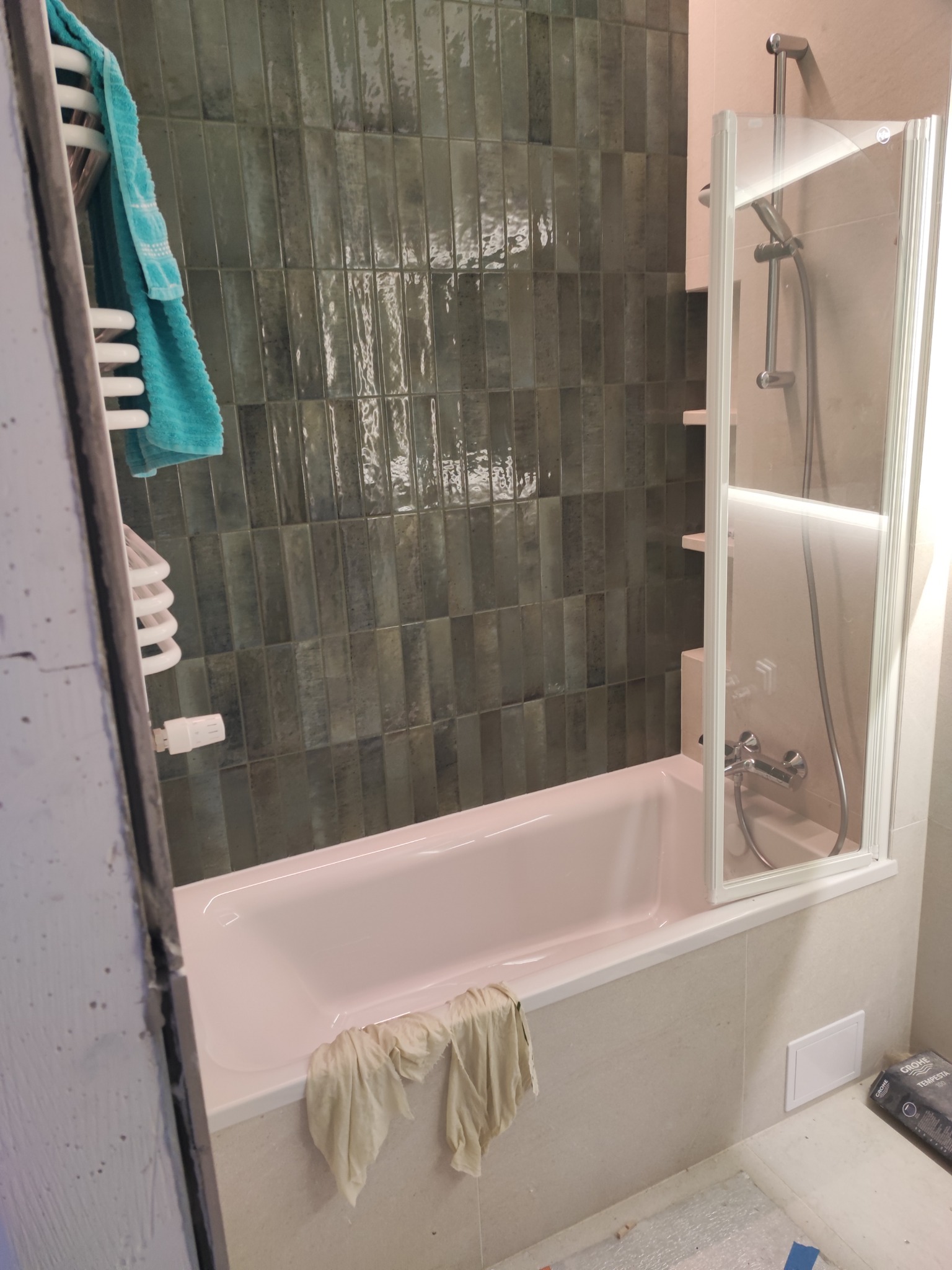 Wykończona łazienka z wanną w odcieniach różu, zielonymi płytkami na ścianie i szklanym parawanem nawannowym. Widoczny grzejnik ręcznikowy i akcesoria łazienkowe.