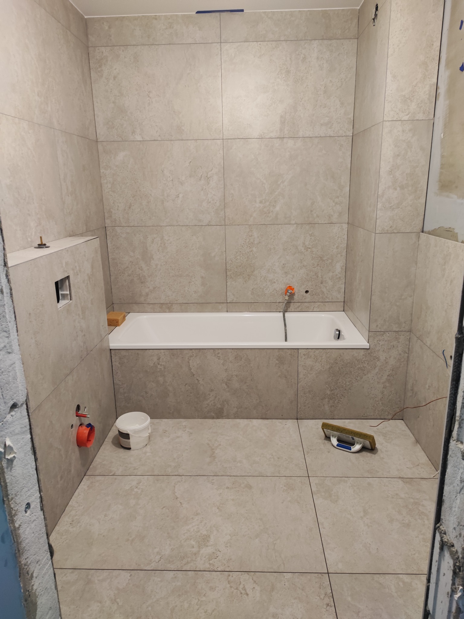 Wykończona łazienka z beżowymi płytkami, białą wanną i akcesoriami. Widoczna fuga, gąbka i wiaderko. Minimalistyczny design, ciepłe oświetlenie.