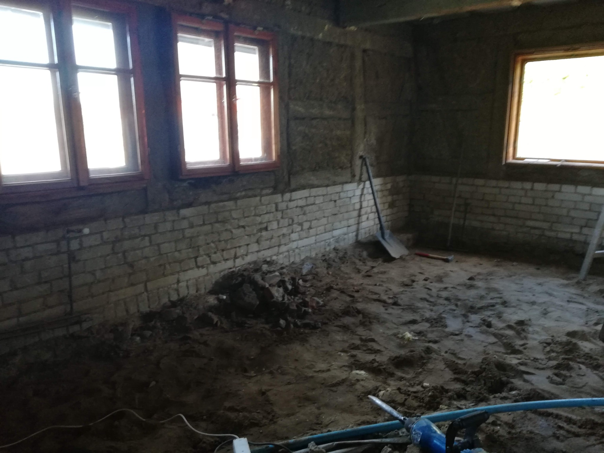 Wnętrze w trakcie prac przygotowawczych: surowe ściany z cegły i izolacji, ziemia na podłodze, narzędzia budowlane. Widoczne okna z drewnianą ramą.