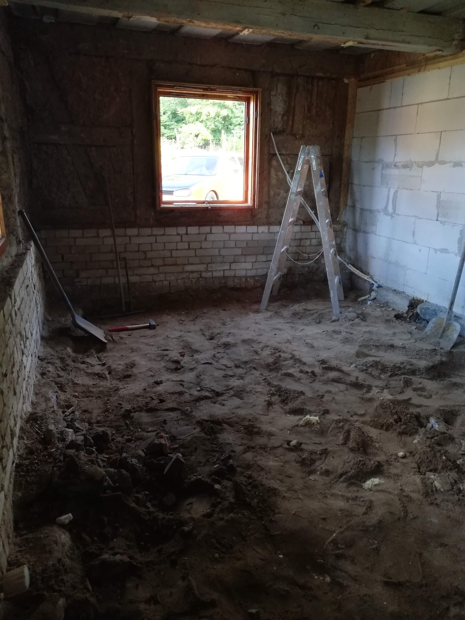 Wnętrze w trakcie prac przygotowawczych do remontu: surowe ściany z cegły i bloczków, ziemia na podłodze, drabina, narzędzia. Widok przez okno na zieleń i samochód.