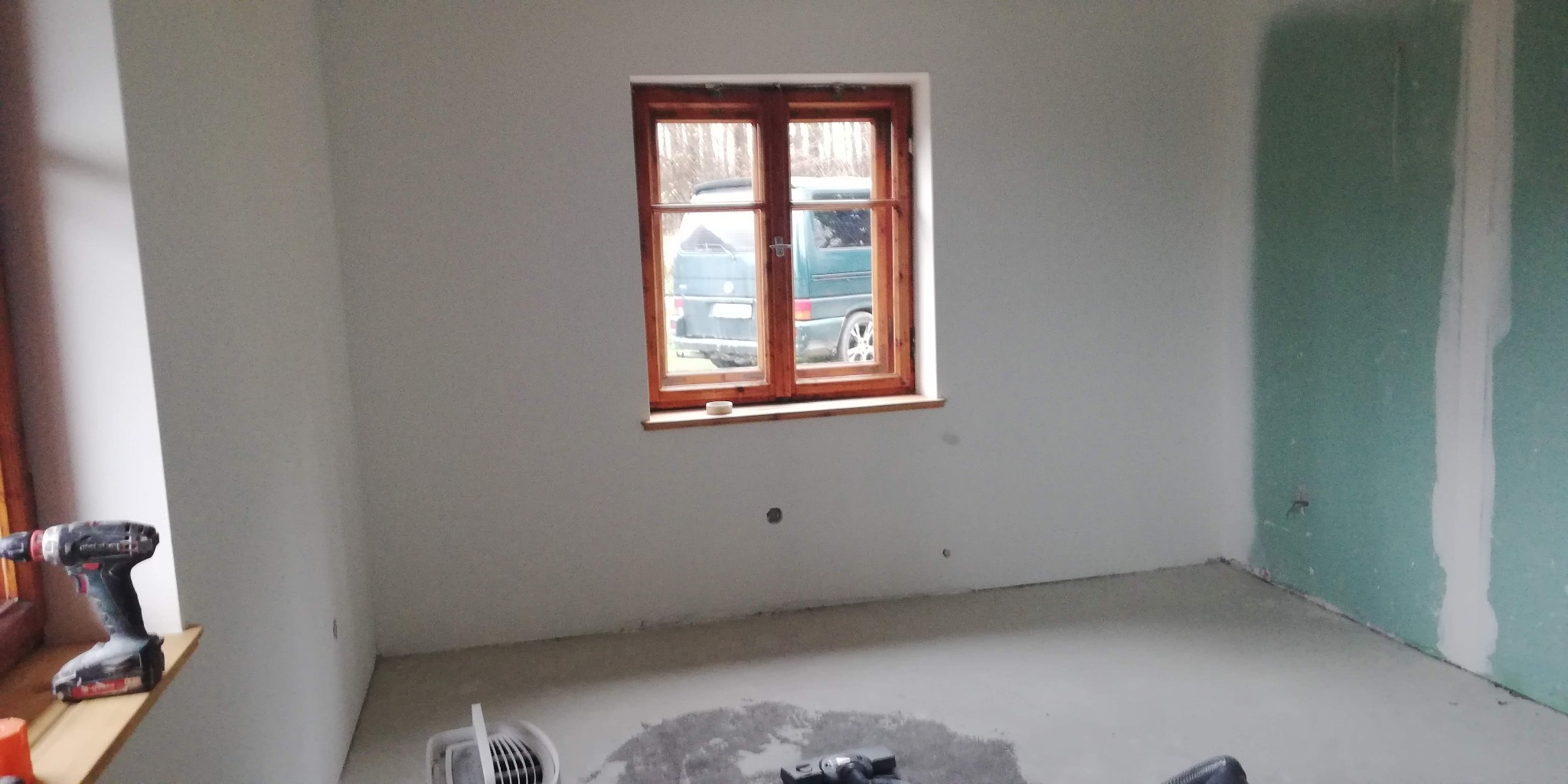 Pokój w trakcie remontu: świeżo położona gładź na ścianach, drewniane okno z widokiem na zewnątrz, narzędzia pracy na parapecie i podłodze. Widoczne ślady prac wykończeniowych.