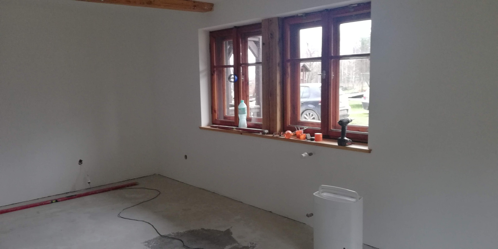 Pokój po gładziowaniu ścian, z drewnianymi oknami i narzędziami na parapecie. Widoczny poziomica na podłodze i biały nawilżacz powietrza. Ściany pomalowane na biało.