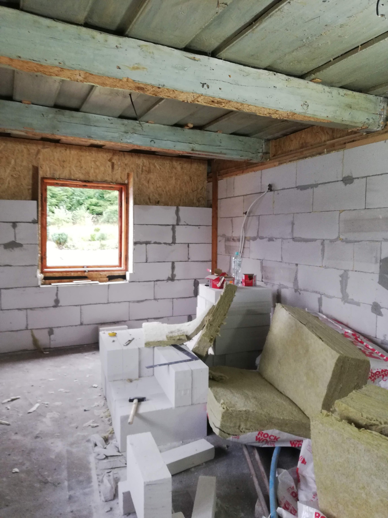 Wnętrze w trakcie budowy: ściany z białych bloczków, drewniane okno z widokiem na zieleń, izolacja z wełny mineralnej i narzędzia budowlane. Surowy stan.
