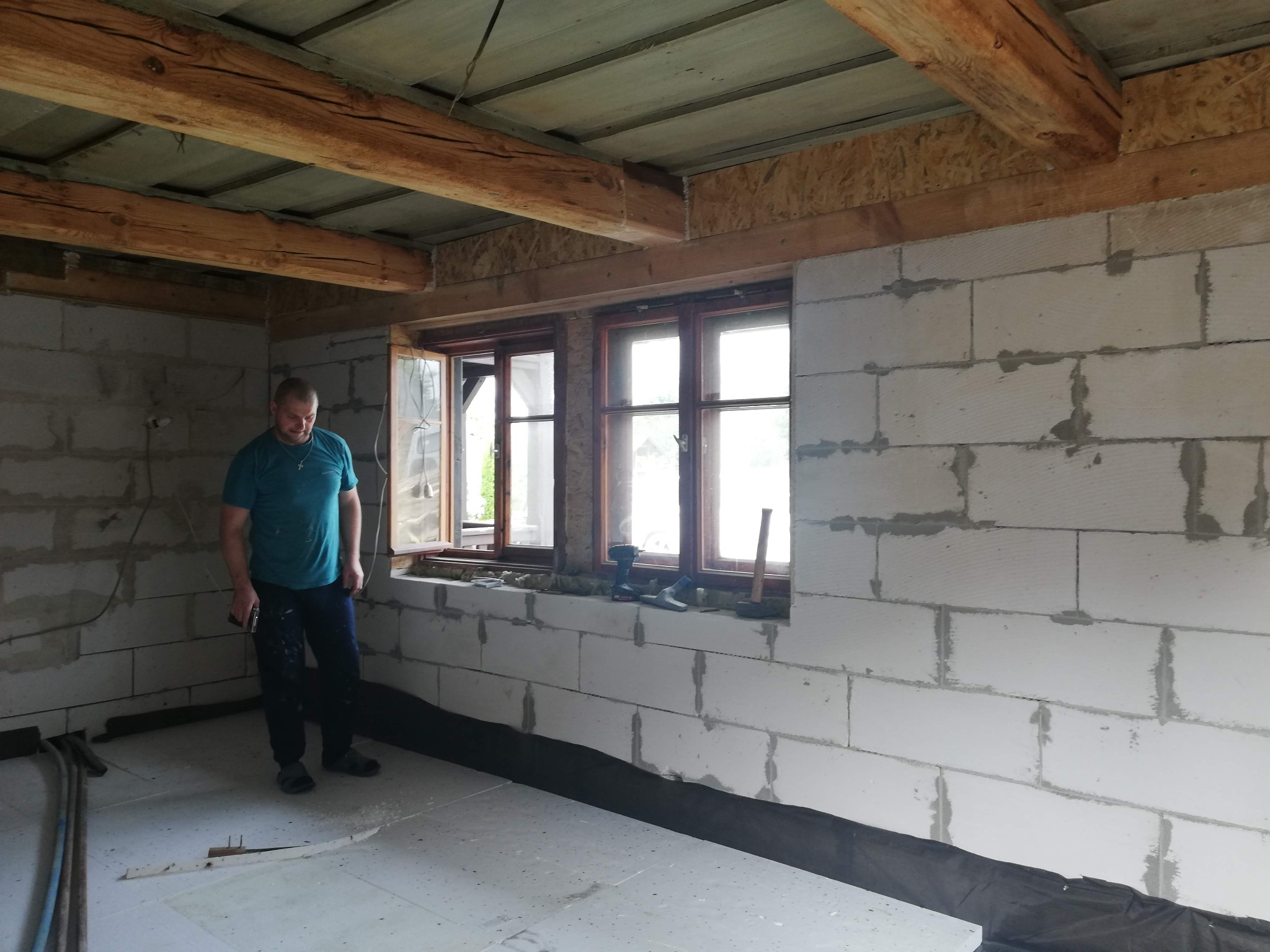 Wnętrze w trakcie prac w Somoninie: ściany z betonu komórkowego, drewniane belki stropowe, okno. Widoczny pracownik i narzędzia.