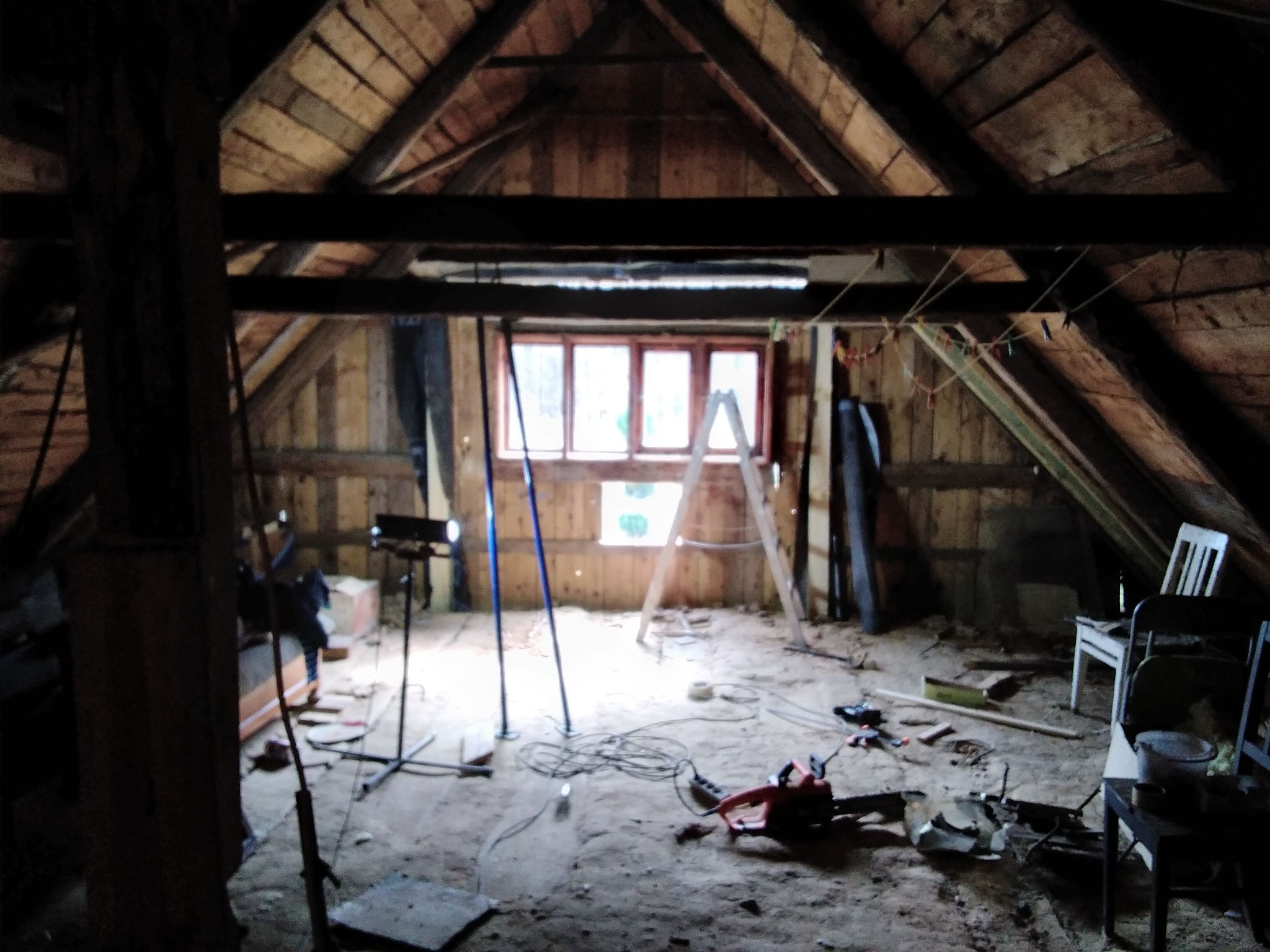 Poddasze w trakcie prac remontowych: drewniana konstrukcja dachu, okno z drabiną, narzędzia i materiały budowlane porozrzucane na podłodze. Widoczne oświetlenie robocze.