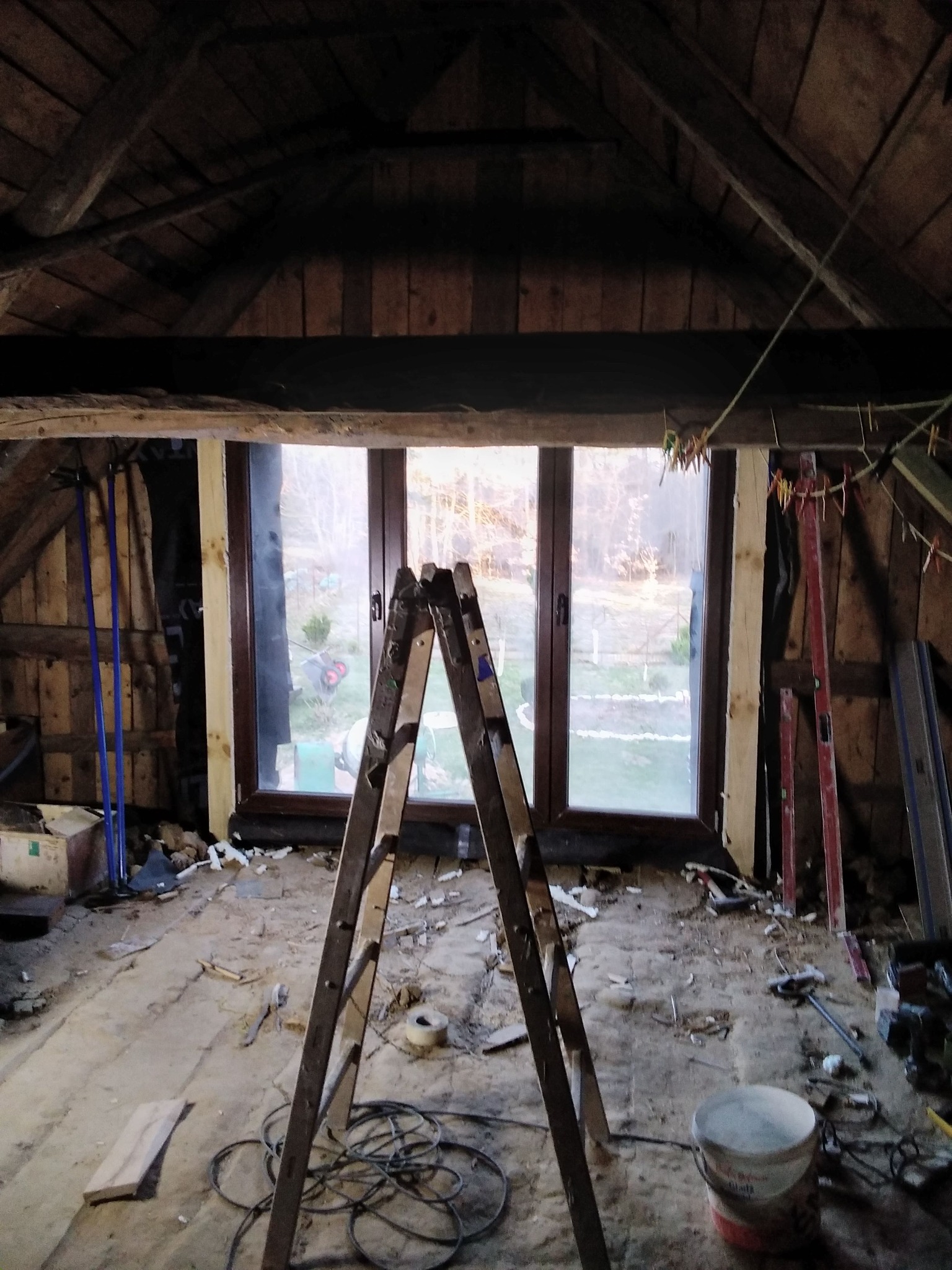 Poddasze w trakcie remontu: drewniana konstrukcja dachu, okno z widokiem na ogród, drabina, narzędzia i materiały budowlane. Widoczny etap prac wykończeniowych.