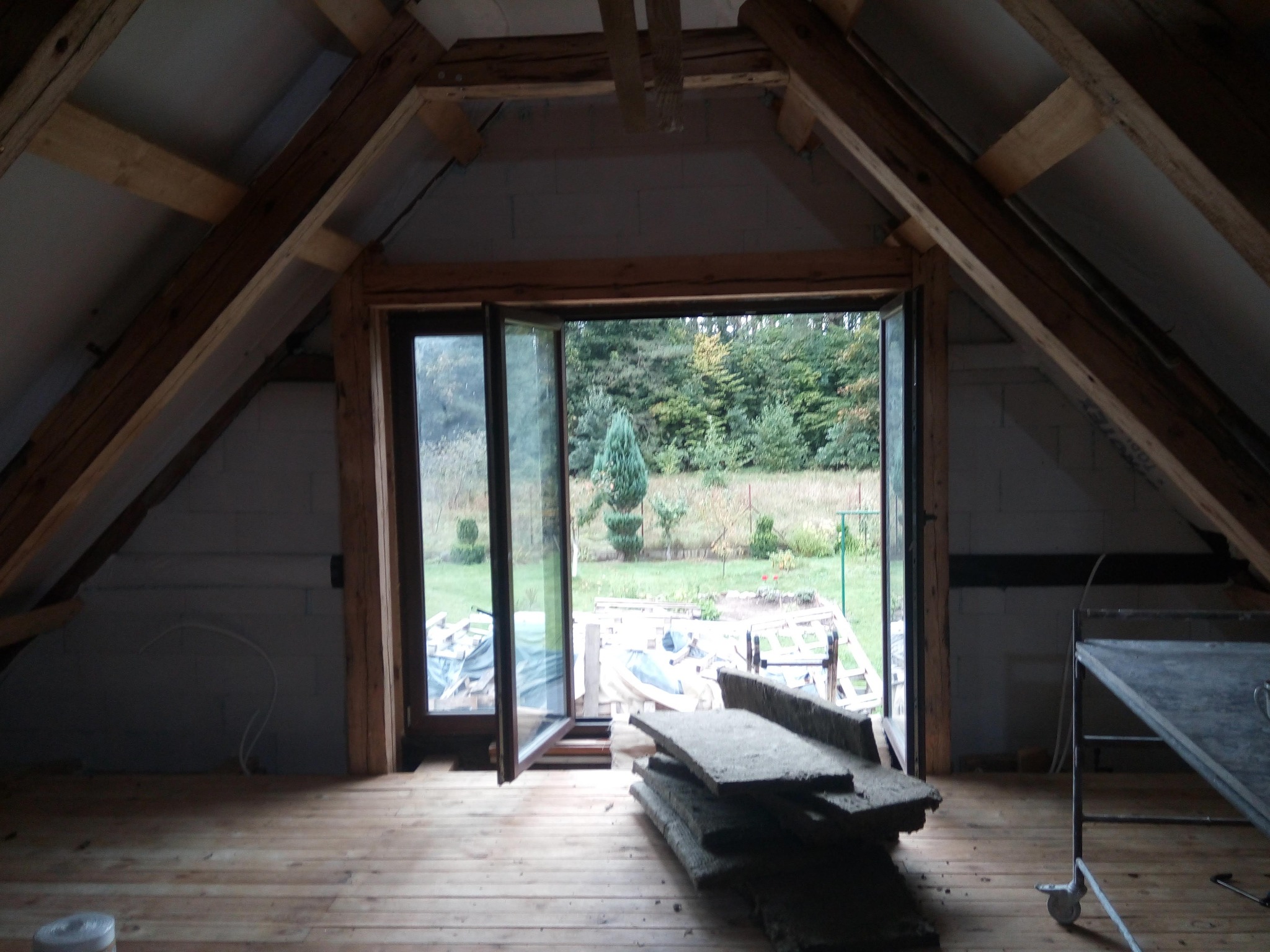 Poddasze z drewnianą konstrukcją, otwartym oknem na ogród i stertą materiałów izolacyjnych na podłodze. Widoczne surowe ściany z bloczków i wózek narzędziowy.