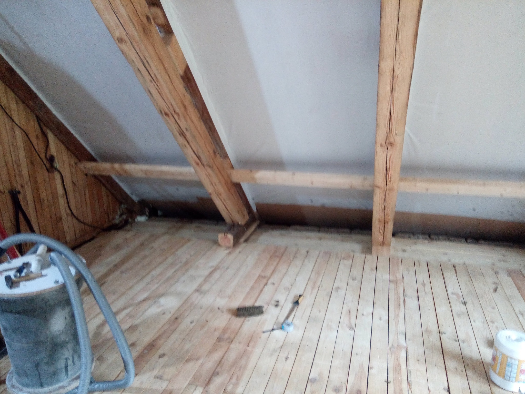 Poddasze z drewnianą podłogą w trakcie prac wykończeniowych. Widoczny odkurzacz przemysłowy, narzędzia i materiały budowlane. Drewniana konstrukcja dachu i skośne ściany.