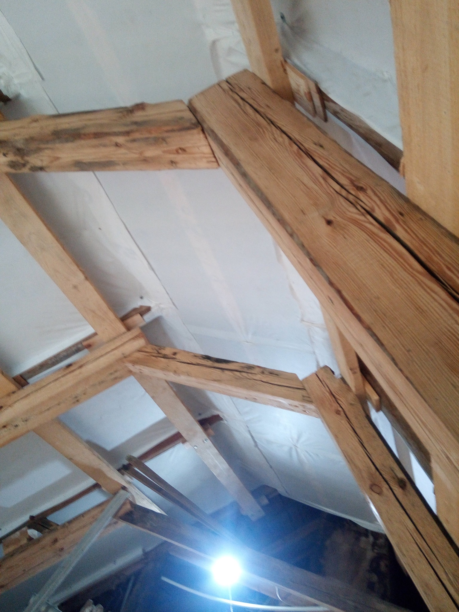 Drewniana konstrukcja dachu z izolacją, widok z dołu. Elementy więźby dachowej z widocznymi słojami drewna i jasną izolacją. Ujęcie z dołu, z silnym źródłem światła.