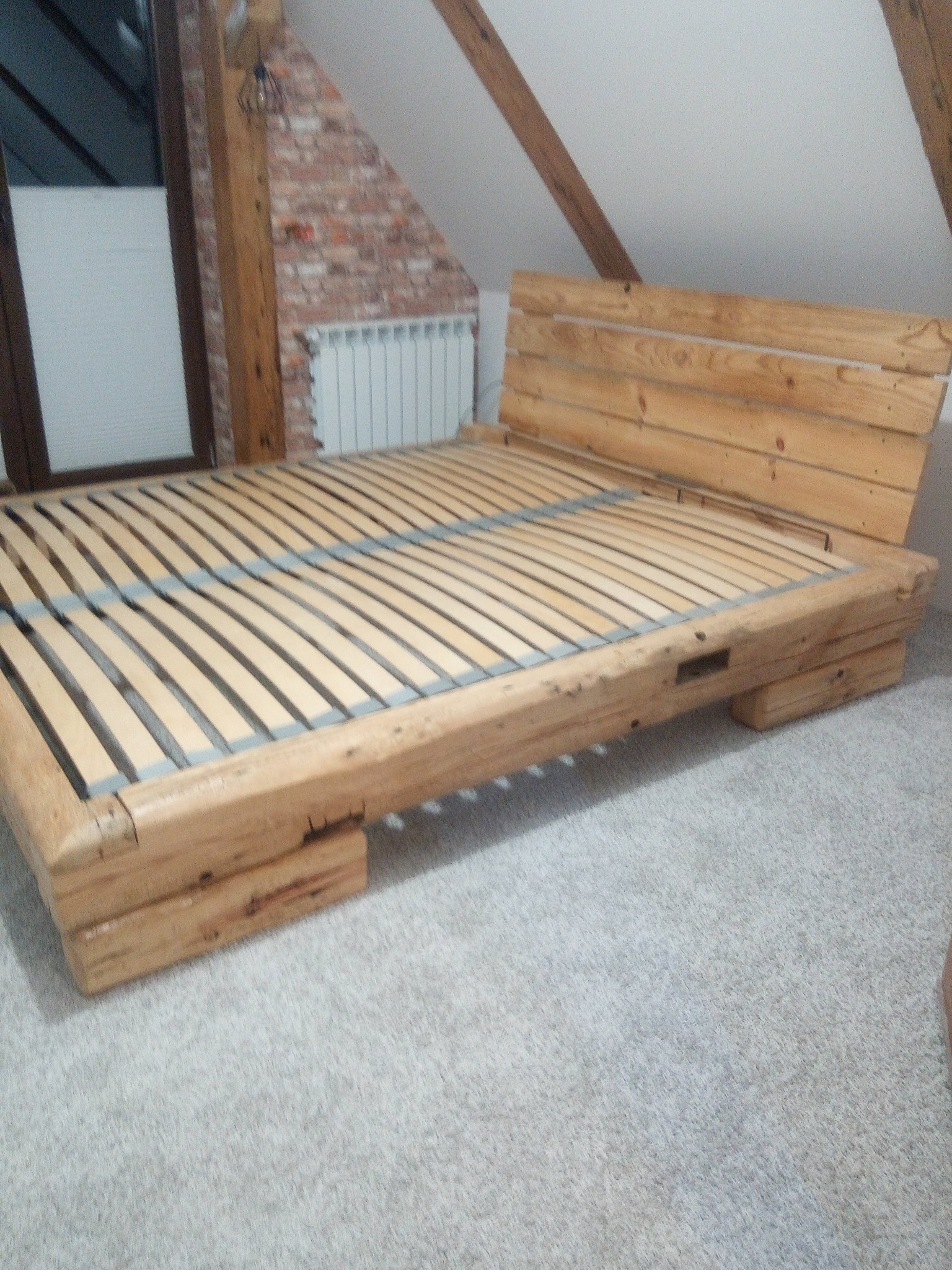 Drewniane łóżko z listwowym stelażem w pokoju na poddaszu. Widoczna ceglana ściana i okno. Naturalne drewno, surowy styl.