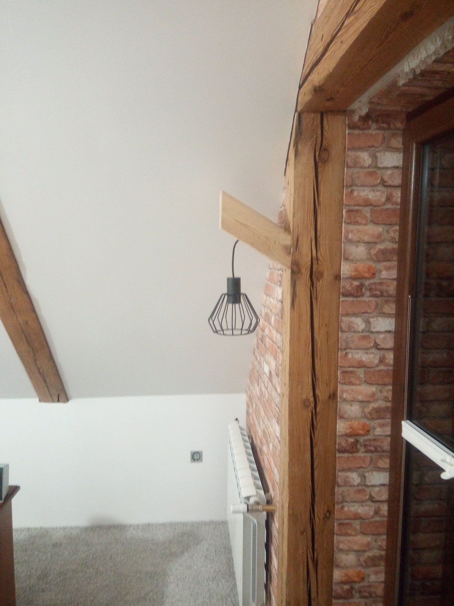 Wykończone wnętrze poddasza z drewnianymi belkami, ceglaną ścianą i nowoczesną lampą wiszącą. Widoczny fragment grzejnika i okna.