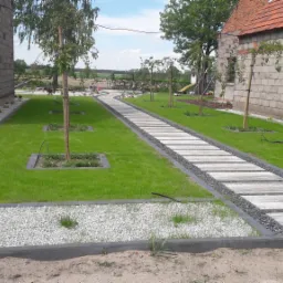 Ścieżka z płyt betonowych prowadząca przez trawnik między budynkami, obrzeża wysypane białym i szarym grysem, młode drzewka w kwadratowych obramowaniach.