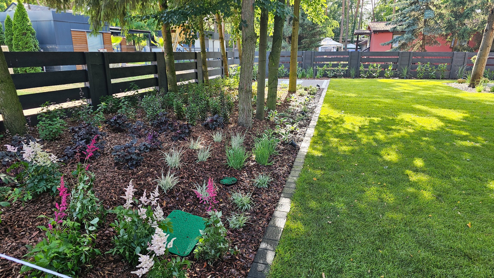 Uporządkowany ogród z rabatami kwiatowymi, drzewami i równo przystrzyżonym trawnikiem, oddzielonym obrzeżem. W tle czarne, drewniane ogrodzenie.