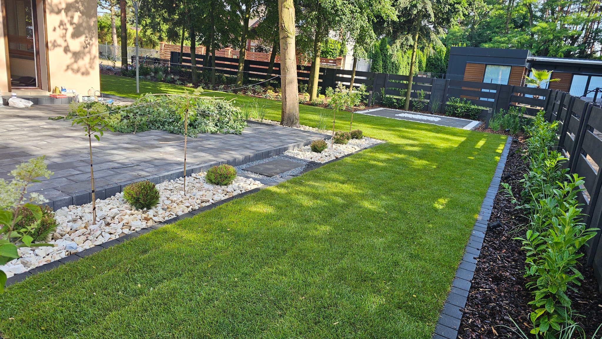 Zadbany ogród z równo przystrzyżonym trawnikiem, ścieżką z kostki brukowej i rabatami z białych kamieni. W tle ciemne ogrodzenie i budynek.