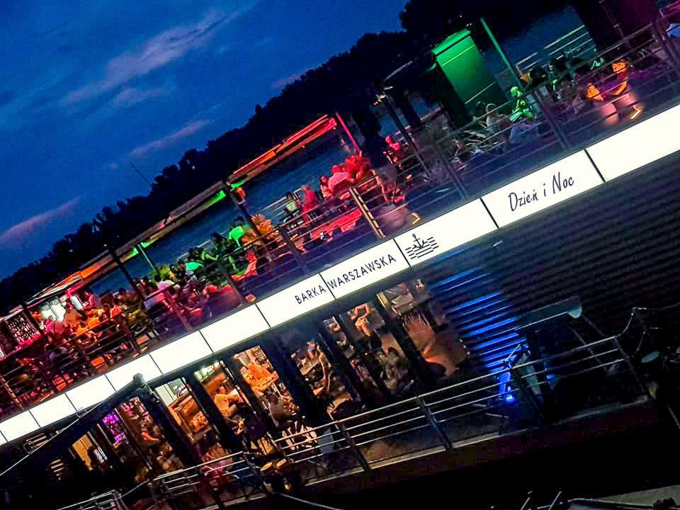 Oświetlona barka 'Barka Warszawska' z napisem 'Dzień i Noc' wieczorową porą, widok z zewnątrz, liczne osoby siedzące przy stolikach.