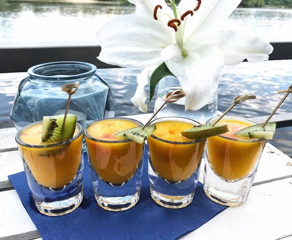 Cztery kieliszki z pomarańczowym napojem, udekorowane kiwi na drewnianych wykałaczkach, ułożone na niebieskiej serwetce na tle białego stołu, z kwiatem lilii i wodą w tle.