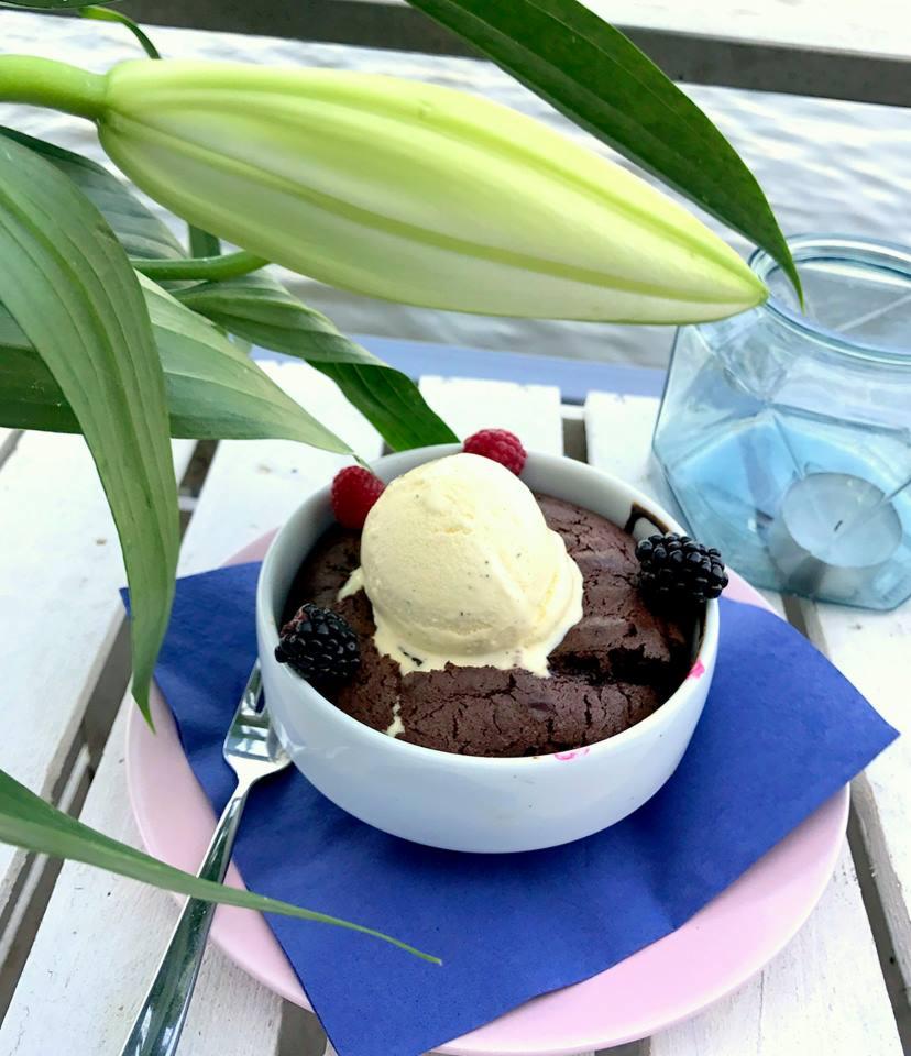 Deser czekoladowy z lodami, malinami i jeżynami w białej miseczce na różowym talerzyku, obok srebrny widelec i kwiat lilii, całość na białym drewnianym stole.