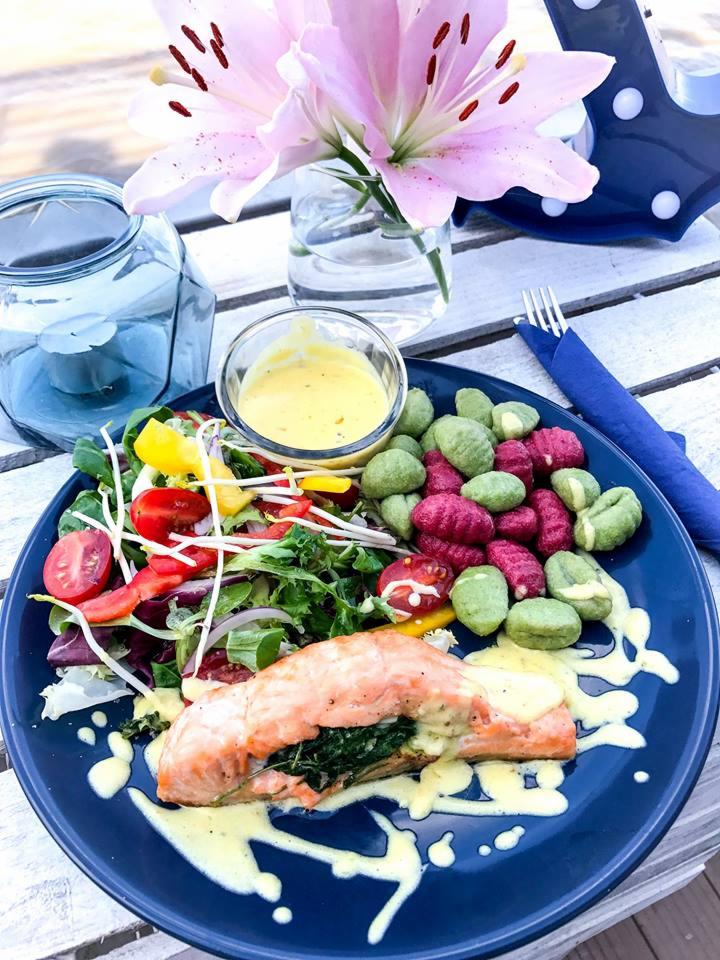 Filet z łososia nadziewany szpinakiem, podany z sosem, sałatką i kolorowymi gnocchi na niebieskim talerzu, udekorowany liliami i świecą.