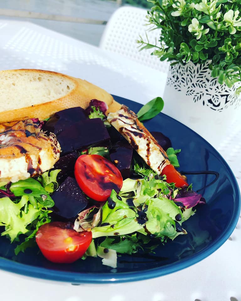 Sałatka z buraków, pomidorów, sera koziego i pieczywa na ciemnoniebieskim talerzu, dekorowana sosem balsamicznym, obok doniczka z zieloną rośliną.