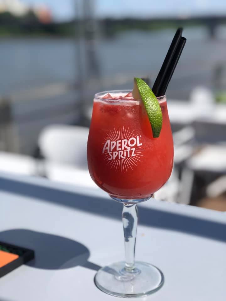 Szklanka Aperol Spritz z lodem i limonką na tle wody, z dwoma czarnymi słomkami.