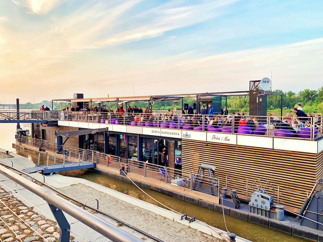 Barka Warszawska zacumowana przy brzegu, z gośćmi siedzącymi na fioletowych pufach na górnym pokładzie, widok z nabrzeża.