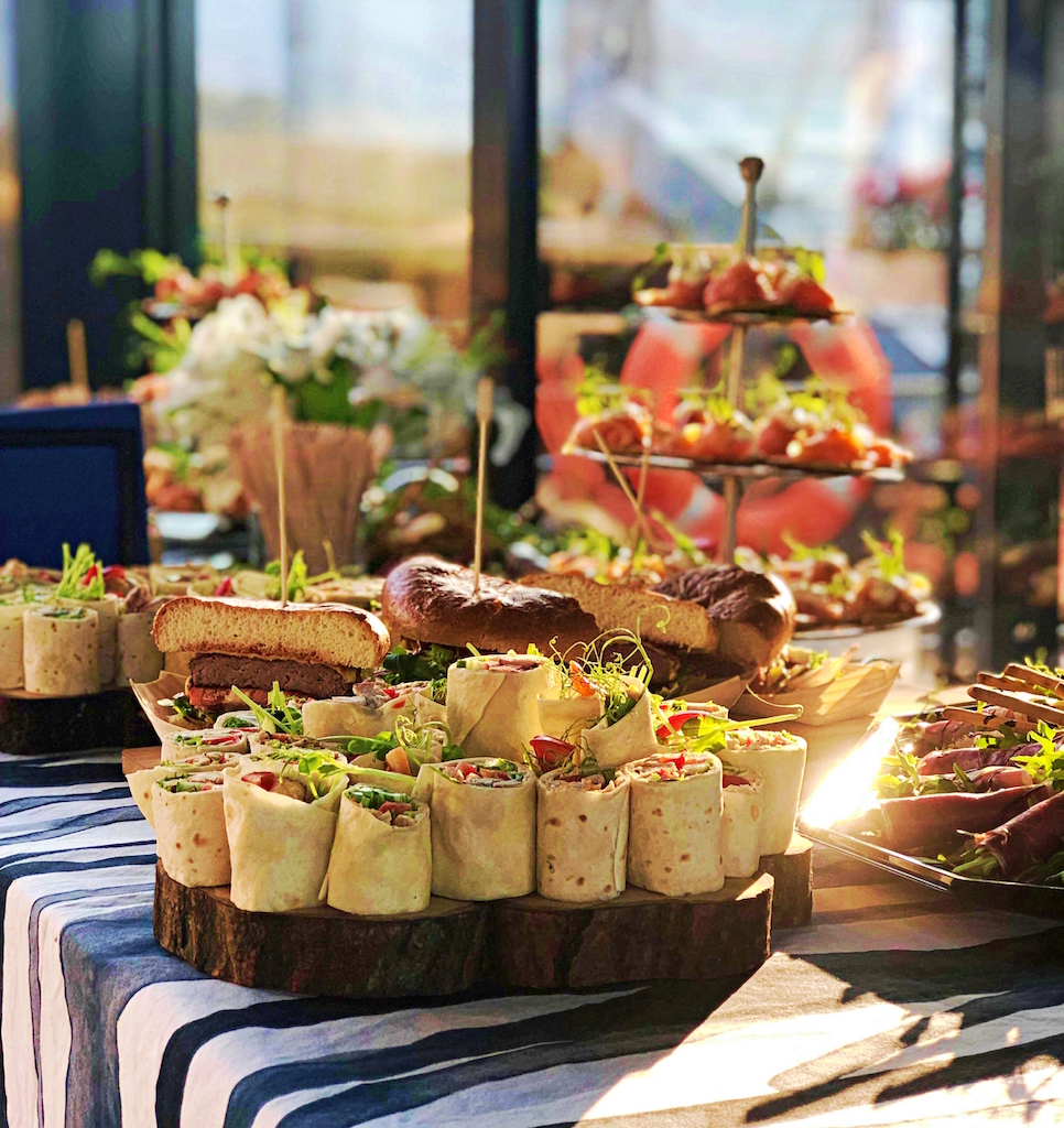 Stół z cateringiem: tortille na plastrach drewna, przekrojone burgery i trzypoziomowa patera z przekąskami, dekoracje kwiatowe w tle, na stole w czarno-białe pasy.