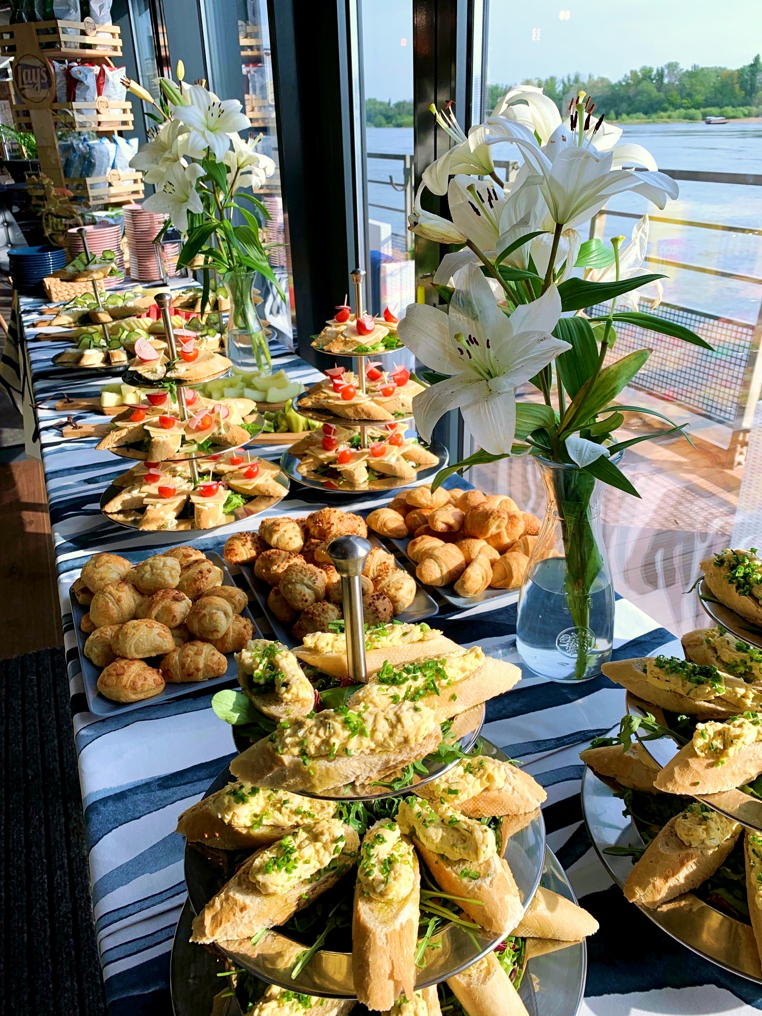 Stół z cateringiem: kanapki z pastą jajeczną posypane szczypiorkiem, mini croissanty, koreczki z pomidorem i serem, białe lilie w wazonie, widok na rzekę zza okna.