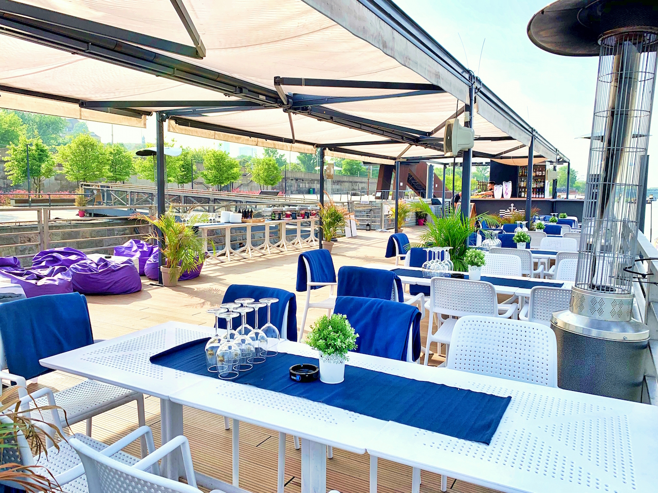 Uroczyste ustawienie stołów z białymi obrusami i niebieskimi bieżnikami, przygotowane na zewnątrz w Warszawie, z fioletowymi pufami w tle i widokiem na rzekę.