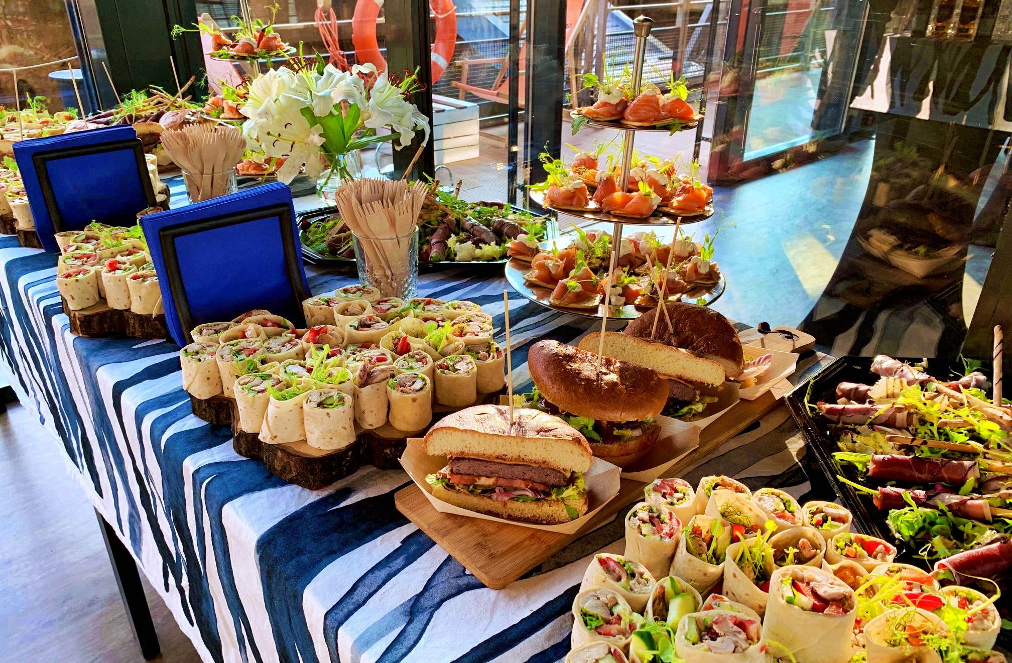 Bogato zastawiony stół z przekąskami: mini tortille na drewnianych podstawkach, burgery przekrojone na pół, roladki z szynki, kanapeczki z łososiem na paterze, kwiaty w wazonie, niebieskie serwetki...