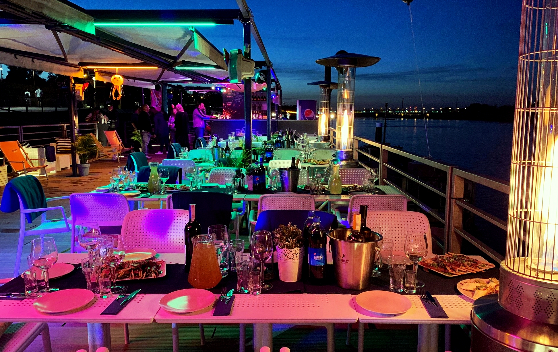 Ustawione stoły na zewnątrz z widokiem na rzekę o zmierzchu, oświetlone kolorowymi światłami, z grzejnikami na gaz i przygotowanym jedzeniem i napojami.