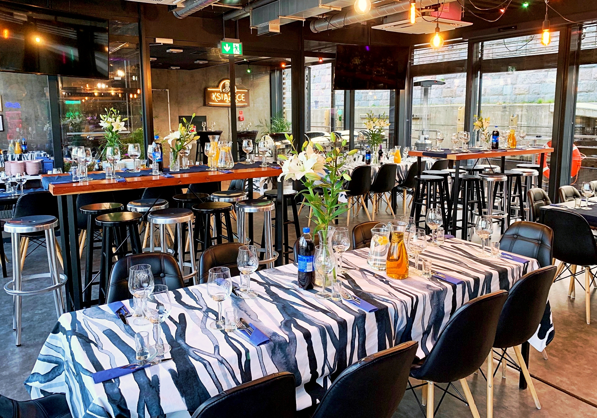 Uroczysta sala restauracyjna z nakrytymi stołami, dekoracjami kwiatowymi, krzesłami w stylu retro i barem w tle.