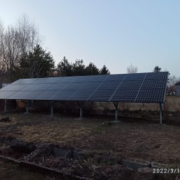 Łukasz Olszak Complex Energy - Rząd paneli słonecznych na metalowych wspornikach w ogrodzie, widoczne drzewa bez liści i budynek gospodarczy w tle, zdjęcie z datą i godziną w prawym dolnym rogu.