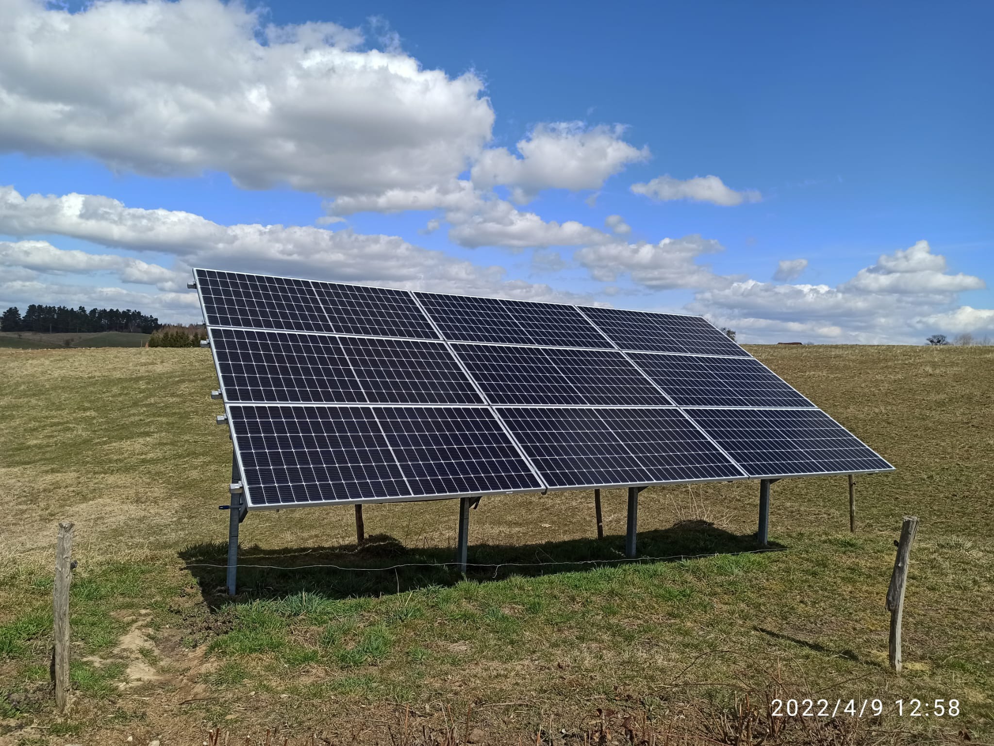 Panel słoneczny ustawiony na łące z widocznymi chmurami na niebie, wsparty na metalowych nogach, otoczony trawą i drewnianym słupkiem ogrodzeniowym.