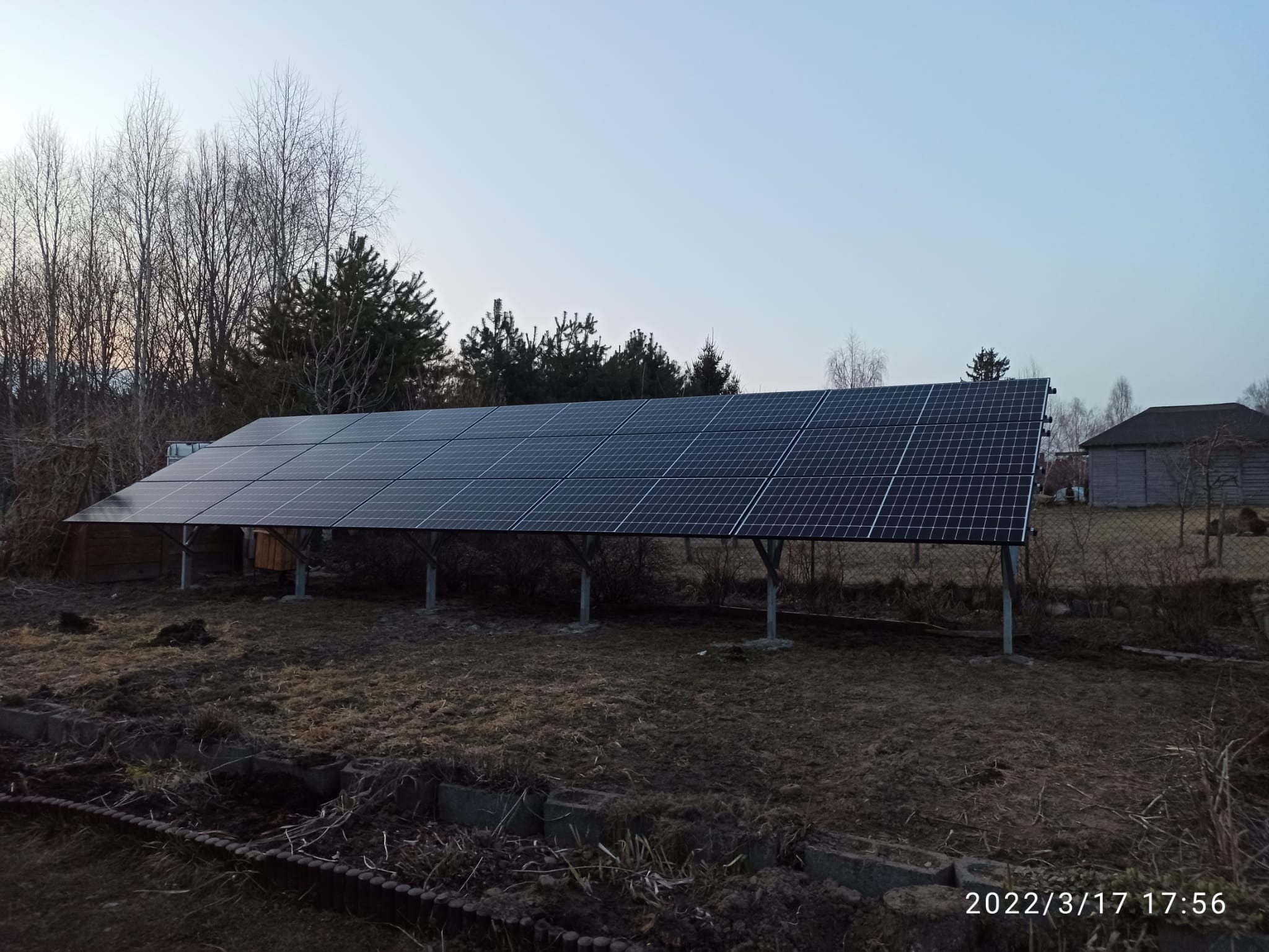 Rząd paneli słonecznych na metalowych wspornikach w ogrodzie, widoczne drzewa bez liści i budynek gospodarczy w tle, zdjęcie z datą i godziną w prawym dolnym rogu.