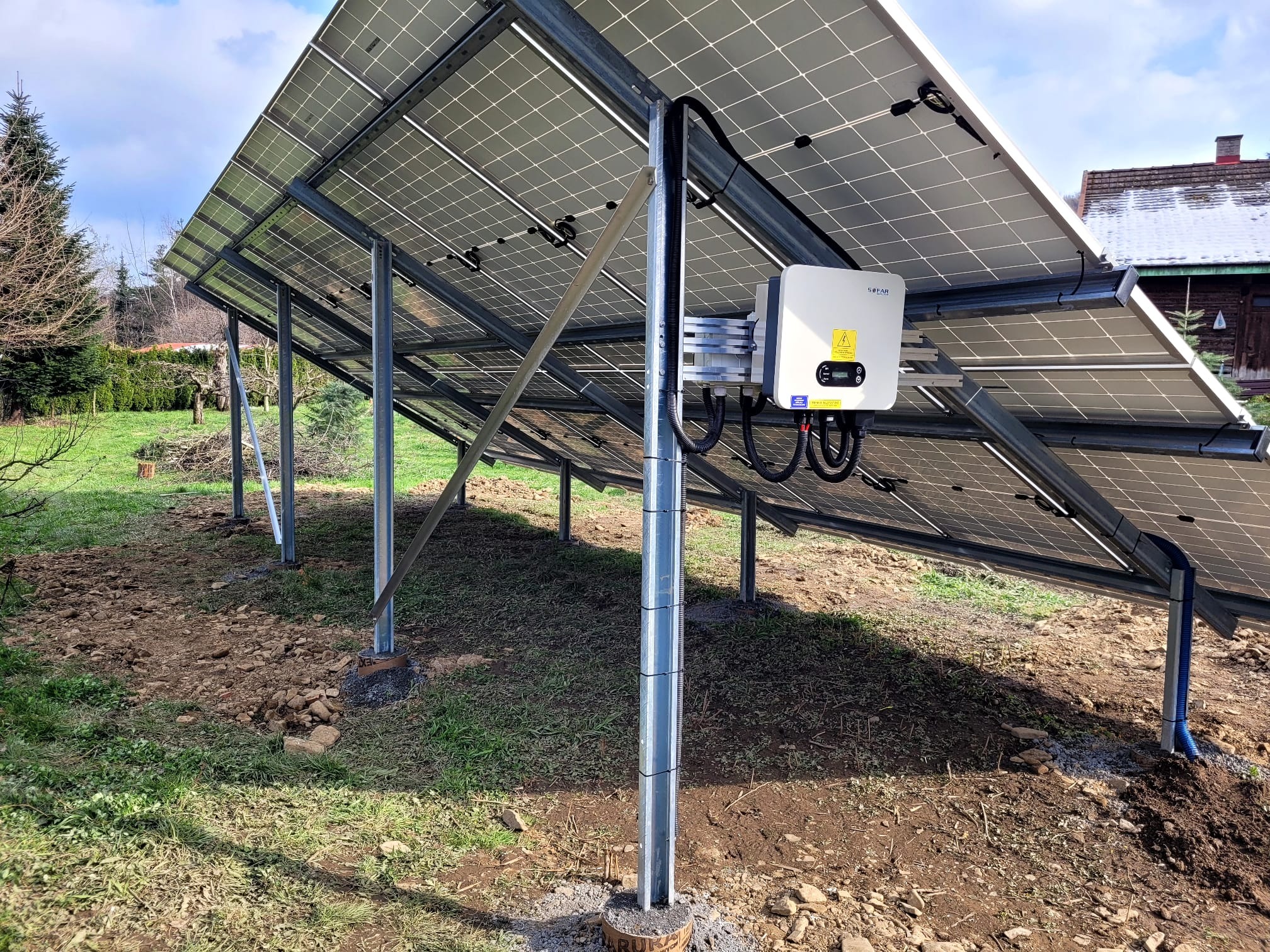 Montaż paneli słonecznych w Olsztynie, widok od spodu na konstrukcję wsporczą i inwerter SOFAR, z widocznymi kablami i elementami mocującymi na tle trawnika i domu.
