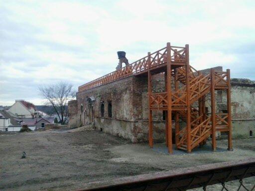 Widok na odrestaurowywany, ceglany budynek z drewnianą konstrukcją schodów i pomostem, na tle szarego nieba i zabudowań miejskich.