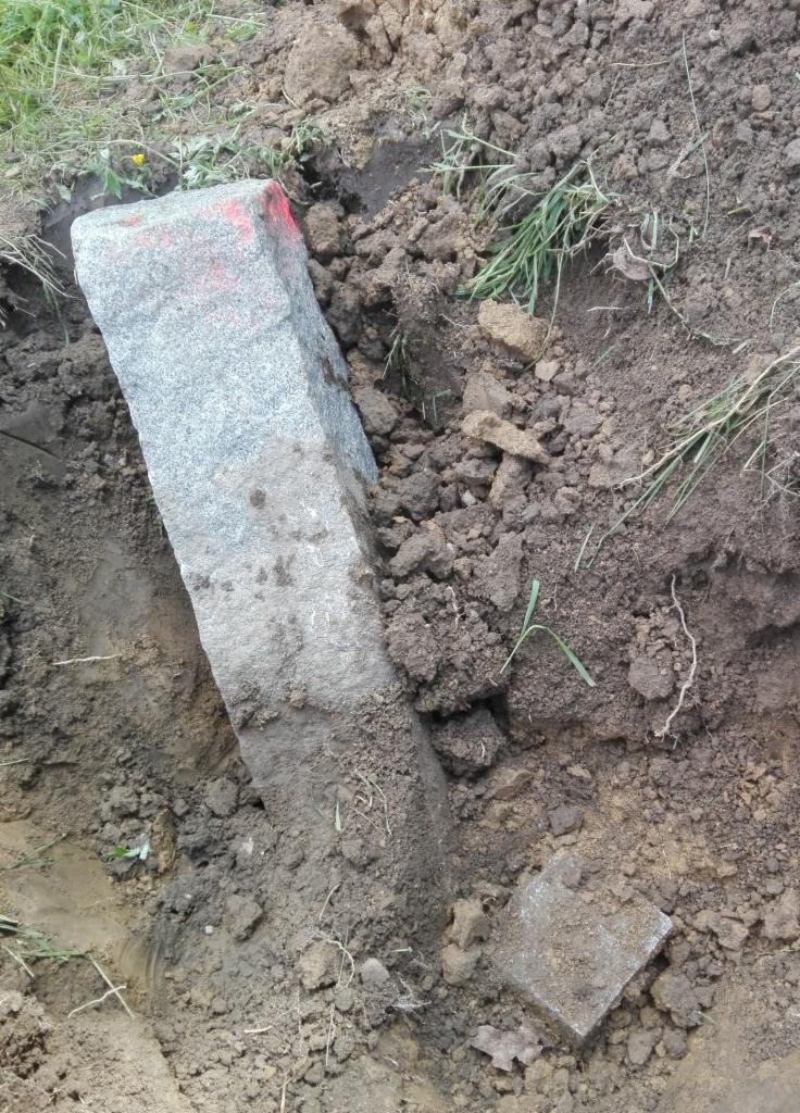 Granitowy obrzeże z czerwoną farbą na krawędzi, częściowo zagłębione w ziemi, otoczone grudami ziemi i źdźbłami trawy. Widoczny fragment mniejszego elementu obrzeża.