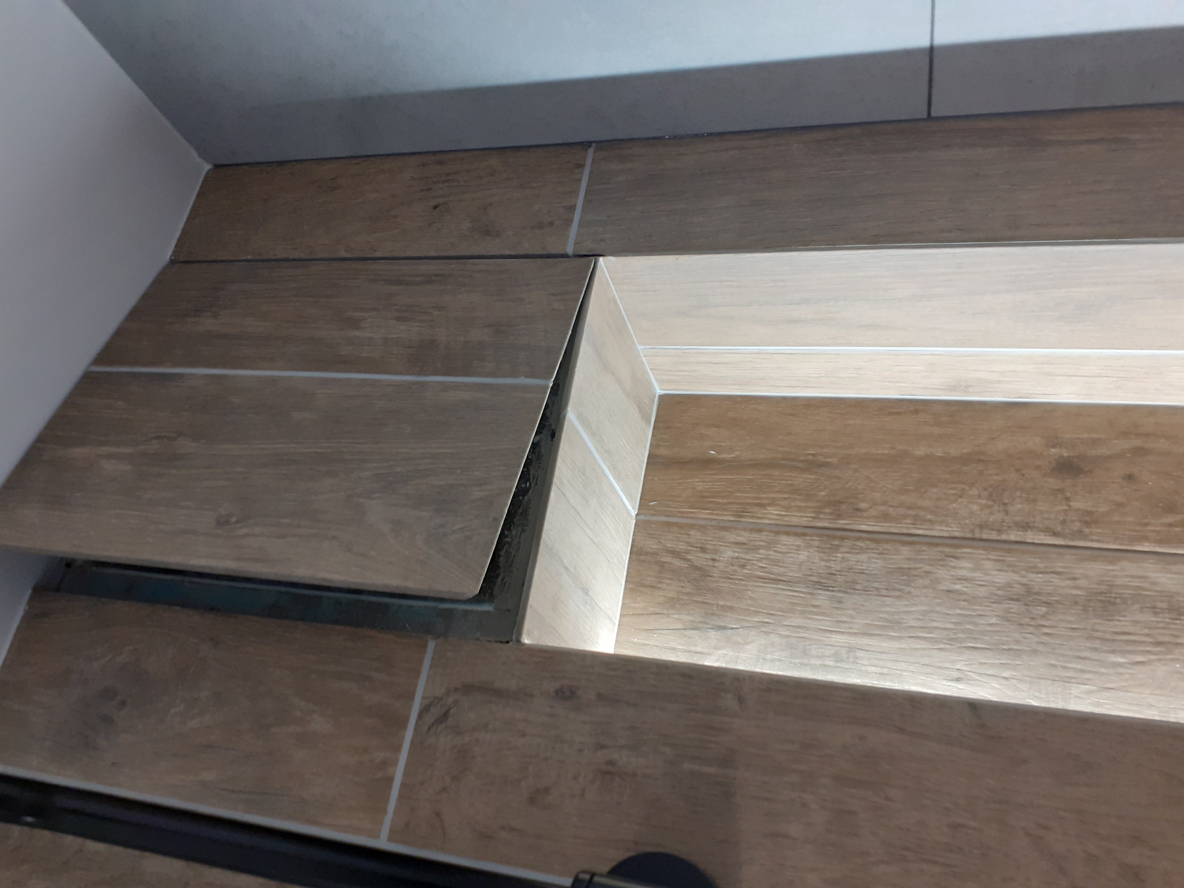 Szczegółowo ułożone płytki imitujące drewno w łazience, z widocznymi fugami i odpływem liniowym w rogu, oświetlone od dołu.