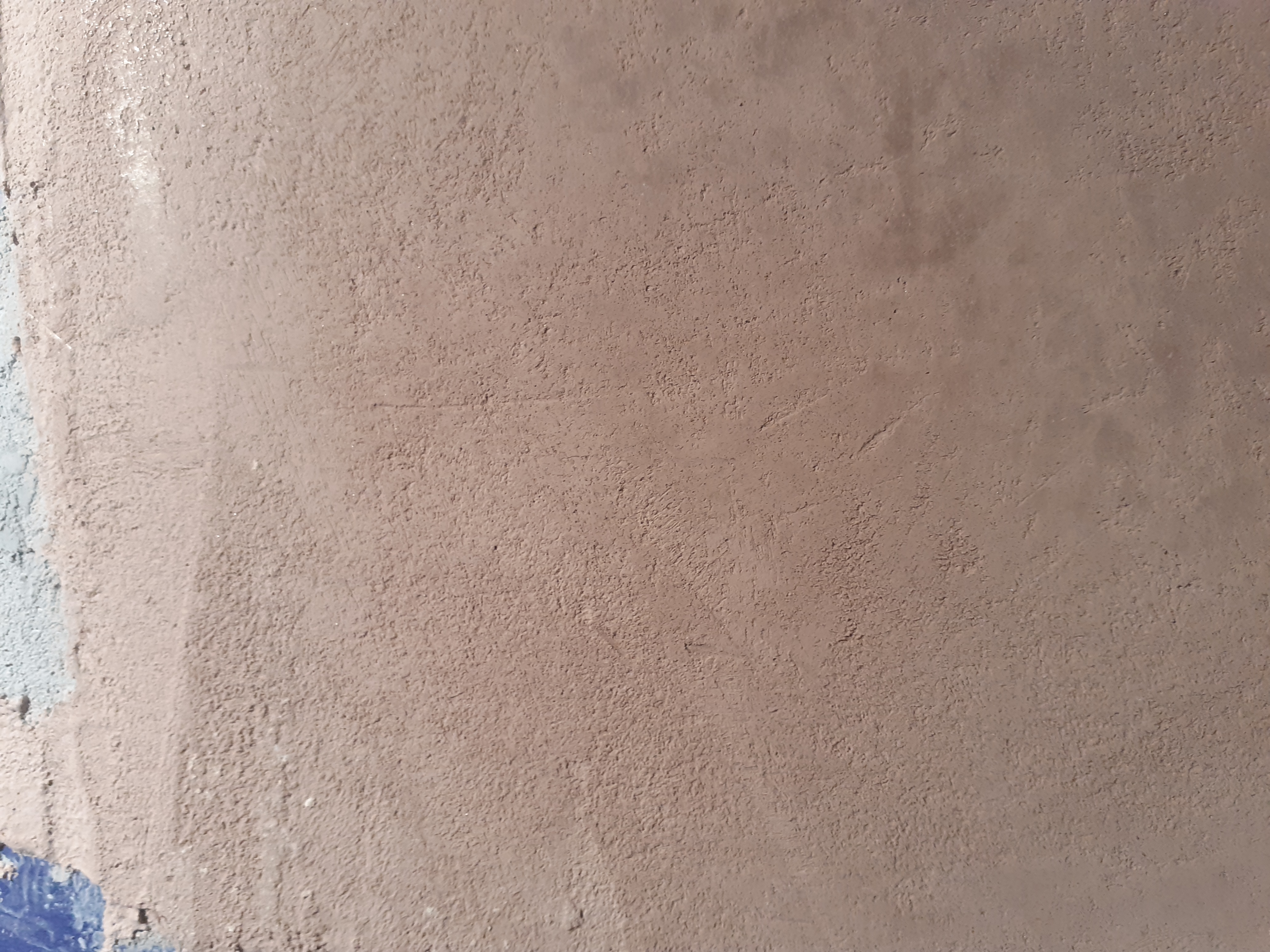 Ściana pokryta świeżo nałożoną, nierówną warstwą gładzi w odcieniach beżu i brązu, z widocznymi śladami narzędzi i fragmentami wcześniejszej, niebieskiej warstwy farby w lewym dolnym rogu.