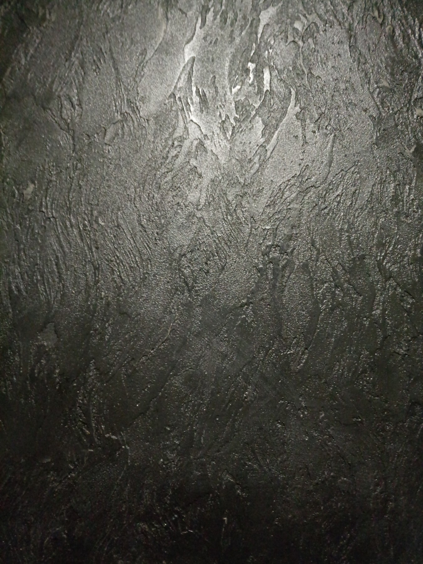Tekstura ściany pokrytej ciemną, metaliczną farbą strukturalną z wyraźnymi nierównościami i refleksami światła.
