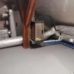 Instalacja systemu w budynku: centrala wentylacyjna z rekuperacją, srebrne rury wentylacyjne na poddaszu, drewniana belka konstrukcyjna, szara podłoga.