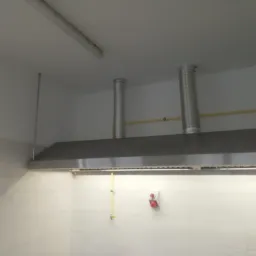 Srebrny okap przemysłowy z trzema kominami wentylacyjnymi w kuchni wyłożonej białymi kafelkami, widoczna instalacja gazowa i wyłącznik bezpieczeństwa.