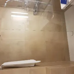 Beżowa kabina prysznicowa z szklaną ścianką, widoczne płytki imitujące kamień, biały ręcznik na siedzisku i chromowana armatura.