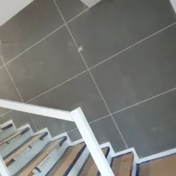 Ściana w szarym kolorze z wyrysowanymi białymi liniami, widoczne schody z białymi barierkami zabezpieczone kartonem i taśmą, ujęcie z góry.