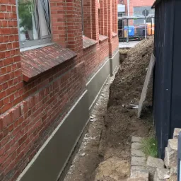 Odkopana ściana fundamentowa z czerwonej cegły, zabezpieczona szarą warstwą izolacji, widoczny wykop i taśma ostrzegawcza.